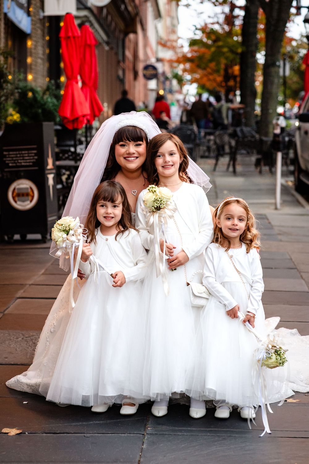 A bridal party in white dresses poses on a city sidewalk during autumn.