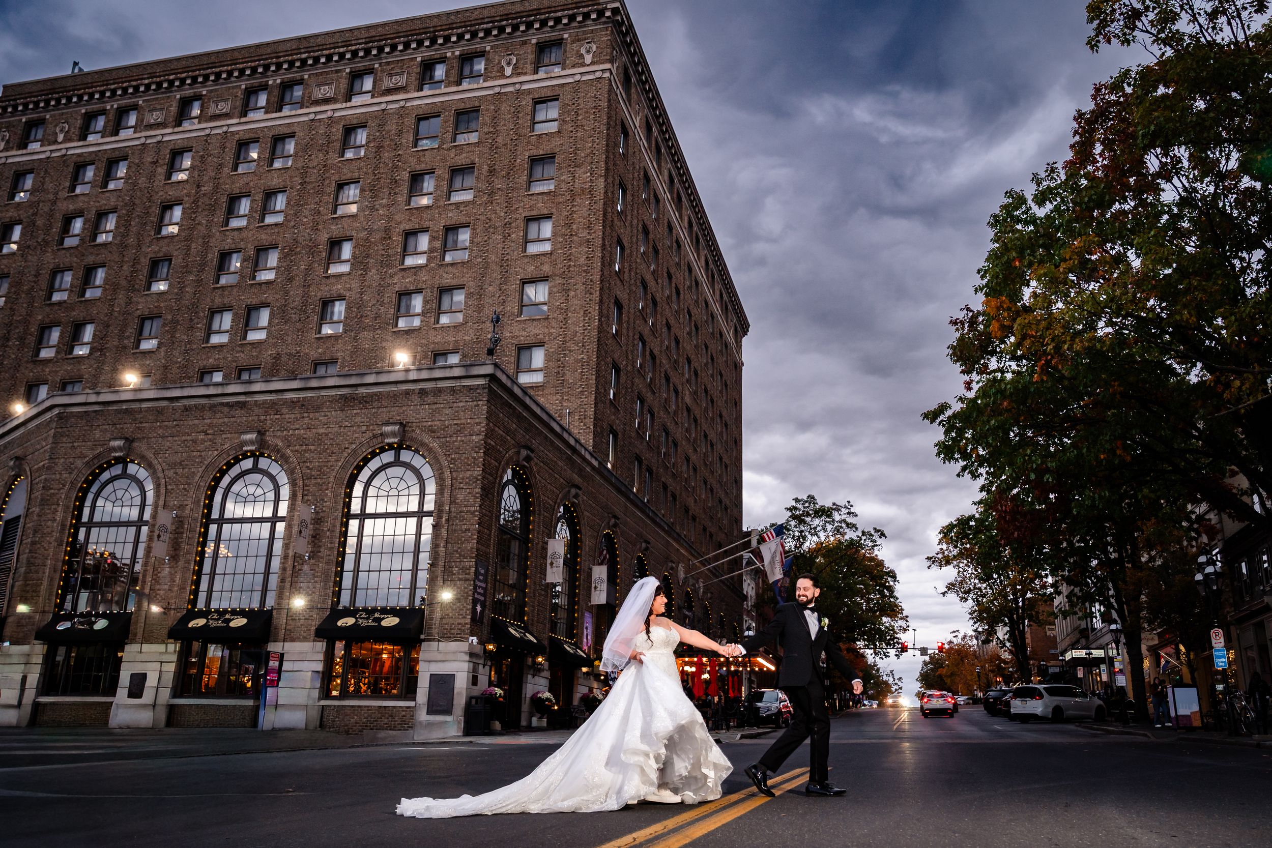 Elegant wedding couple poses outside historic brick apartment building at dusk with dramatic cloudy sky.