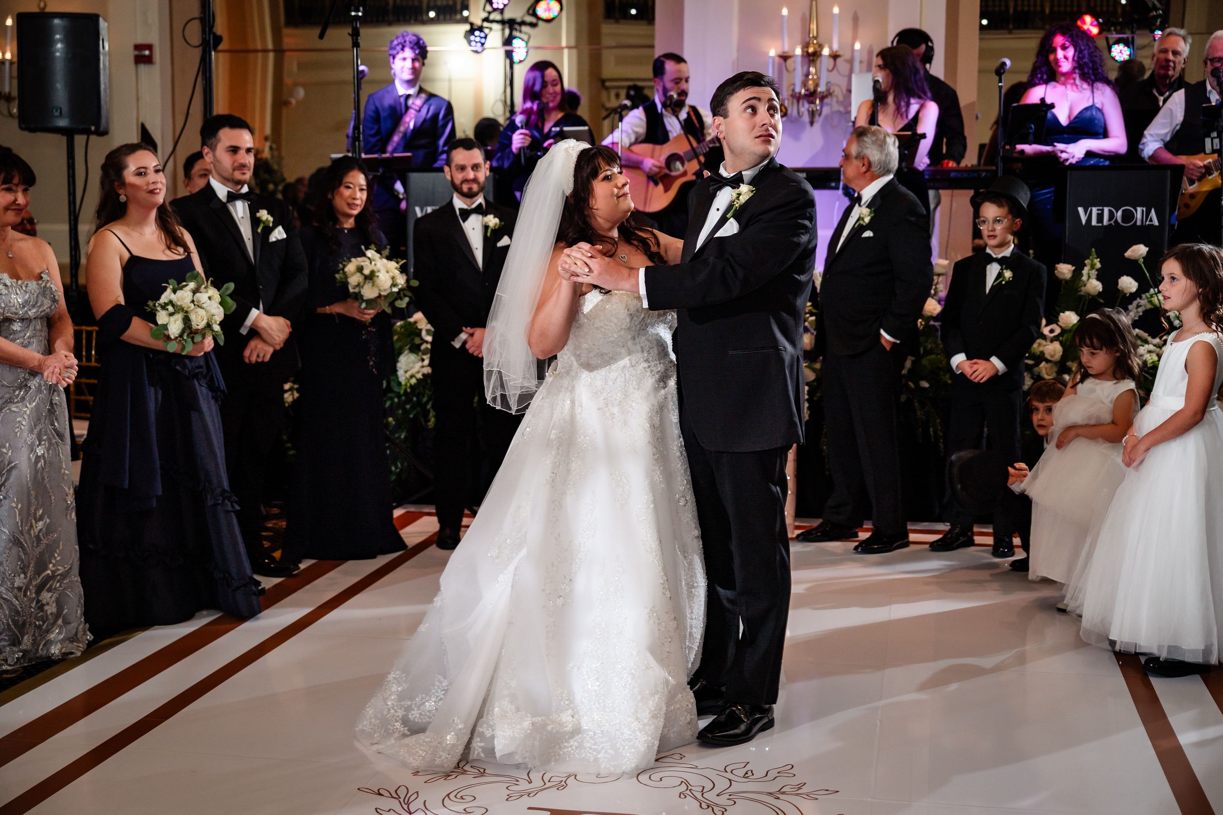 Wedding guests dance and celebrate on an elegant ballroom floor illuminated by purple lighting.