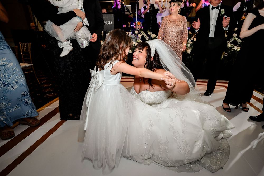 A bride in a white wedding dress hugs a young flower girl during a reception celebration.