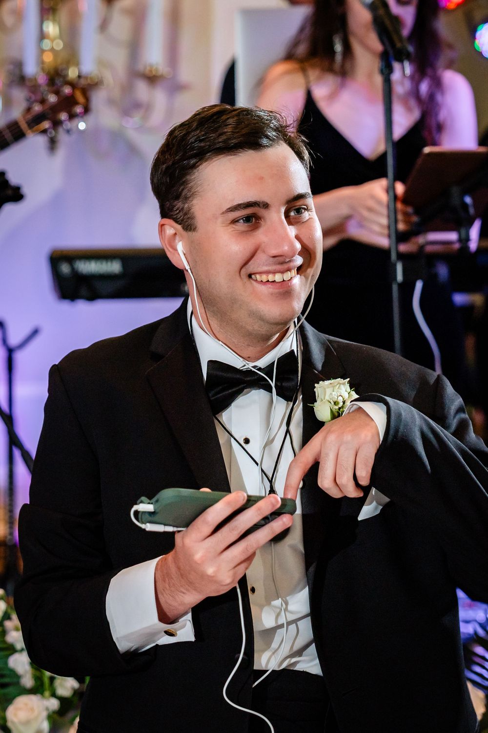Groom in black tuxedo smiling while holding phone during wedding reception.