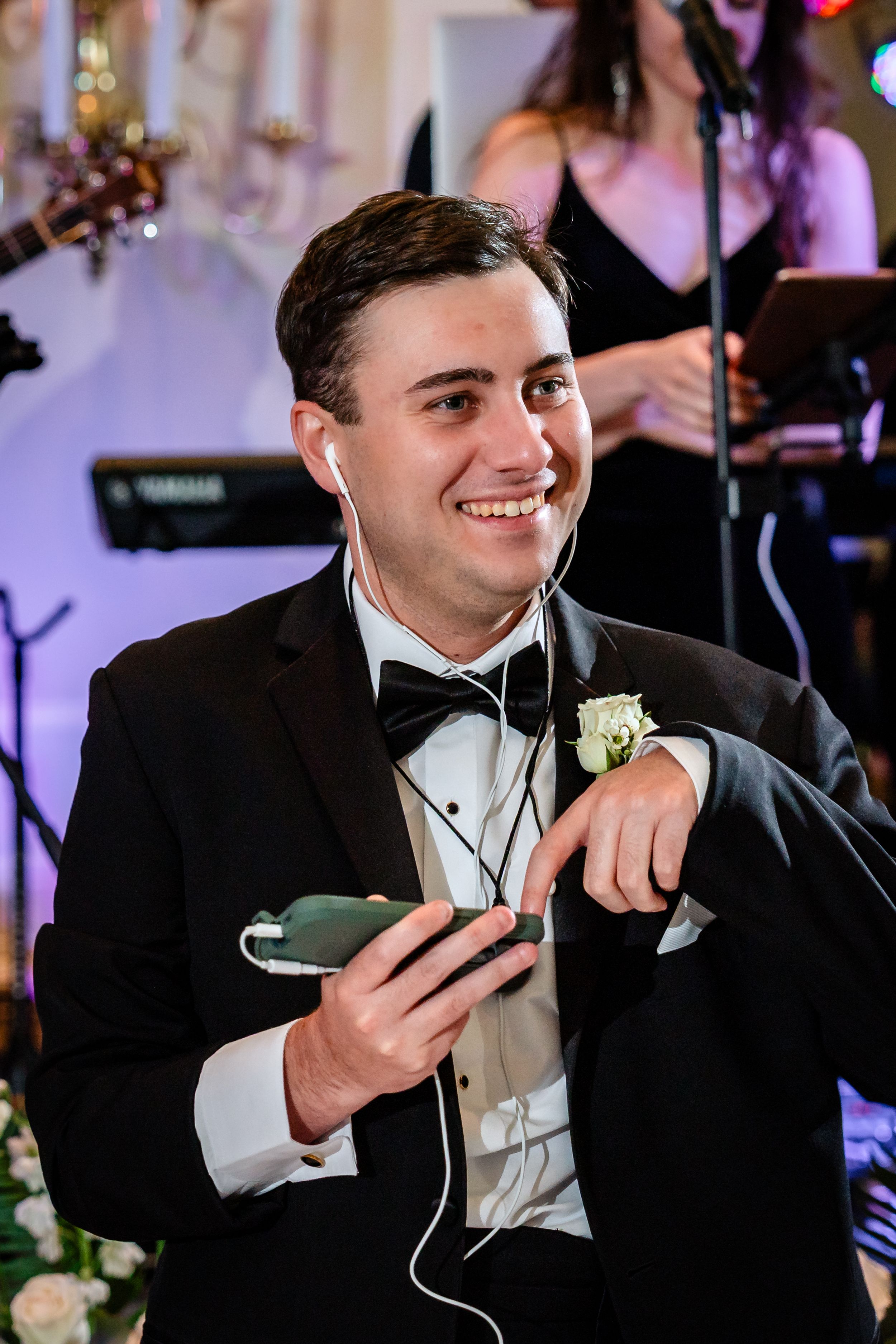 Groom in black tuxedo smiling while holding phone during wedding reception.