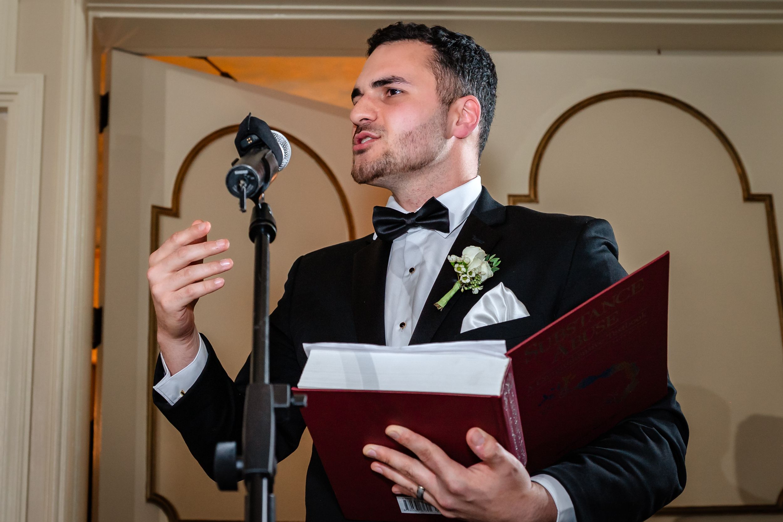 Groom reading from burgundy folder during wedding ceremony in elegant indoor venue.