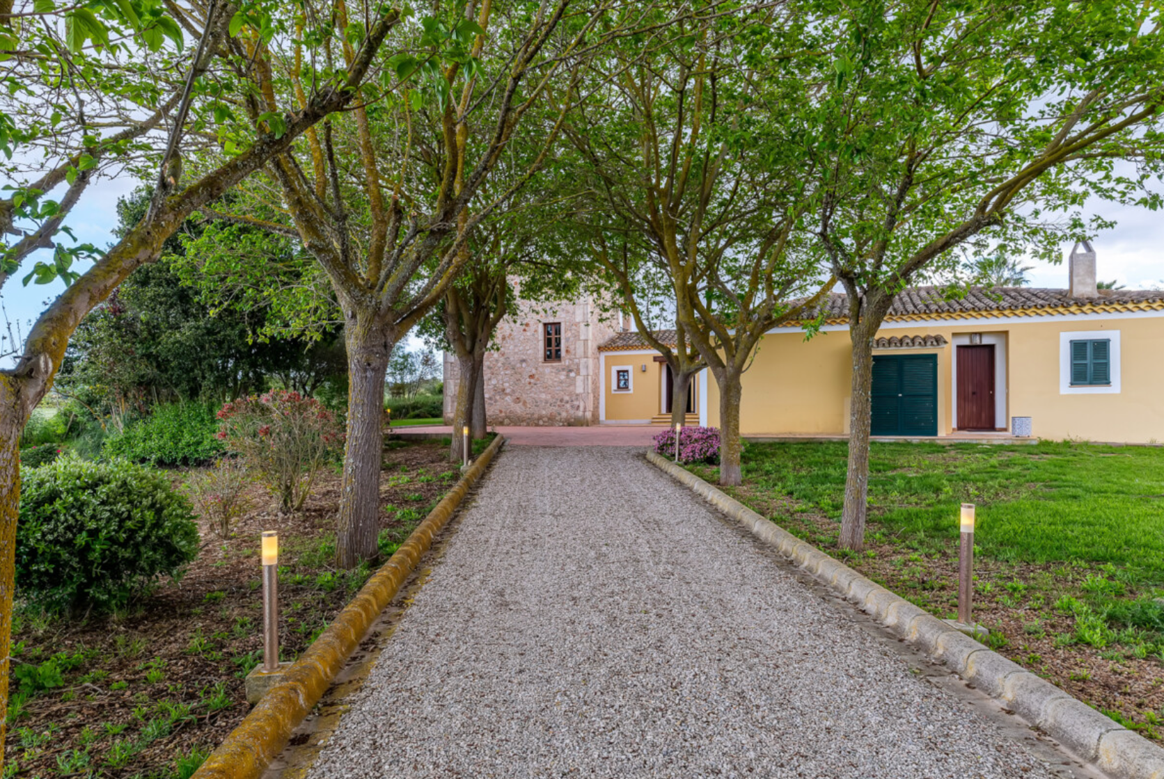 A winding cobblestone pathway leads through a lush garden with stone walls and historic buildings on either side.