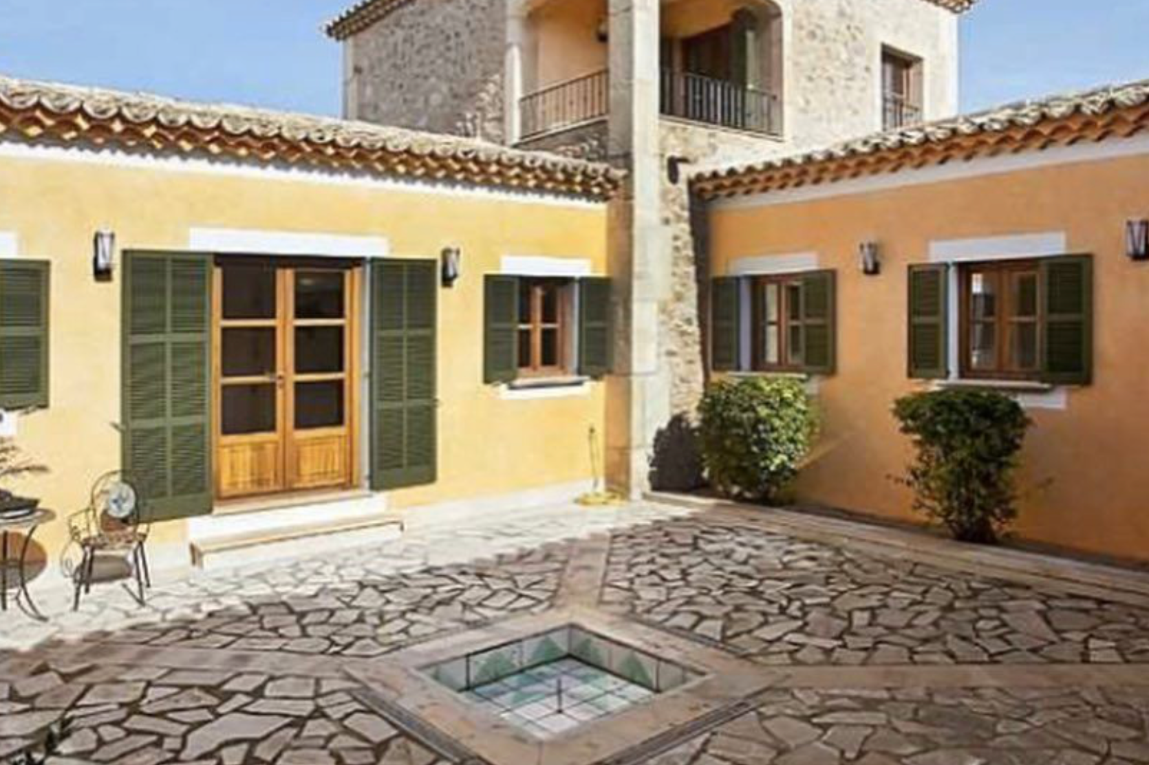 Stone courtyard with rustic paving surrounds a modern villa featuring green shutters and warm terracotta walls.