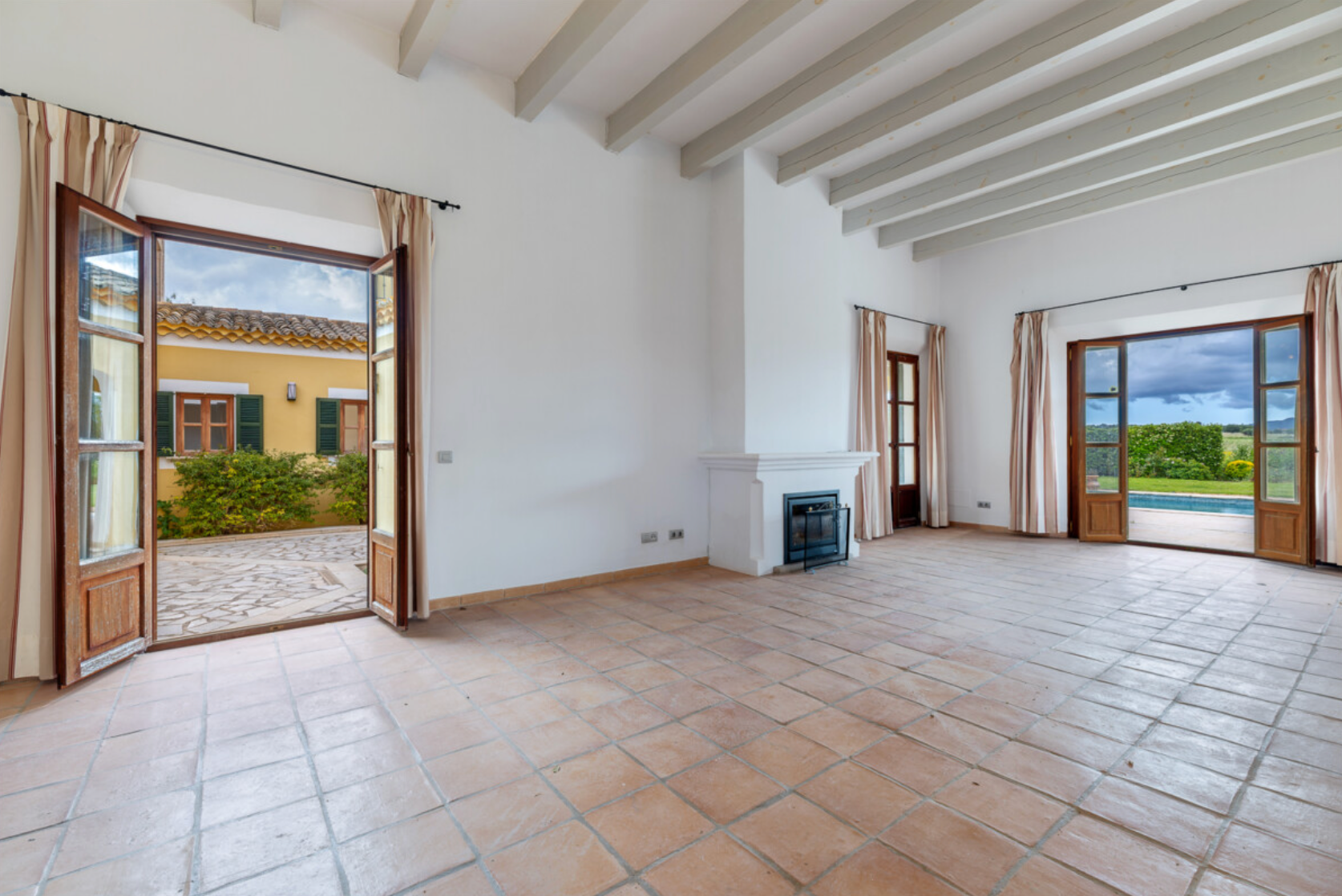 Large empty room with white beamed ceiling, terracotta tile floors, and panoramic windows overlooking scenic countryside views.