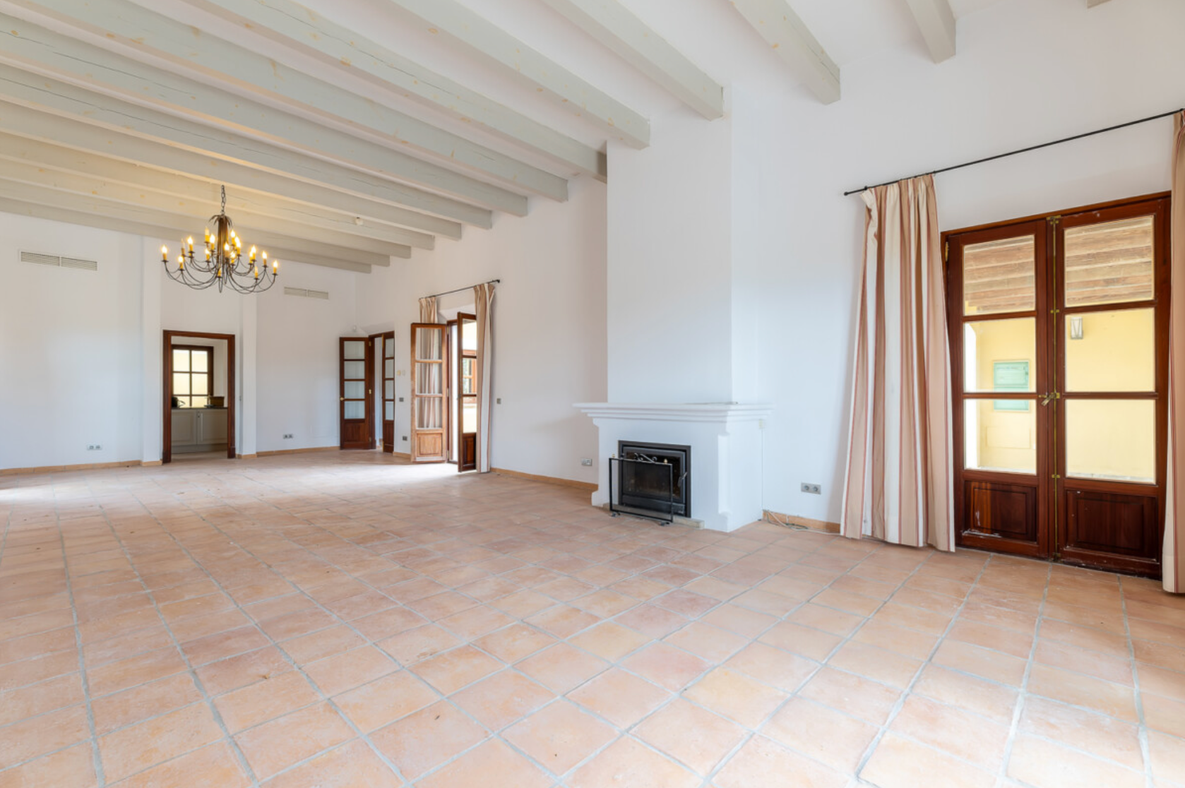 Spacious empty room with white walls, exposed wooden beams, terracotta floors, French doors and a small fireplace.