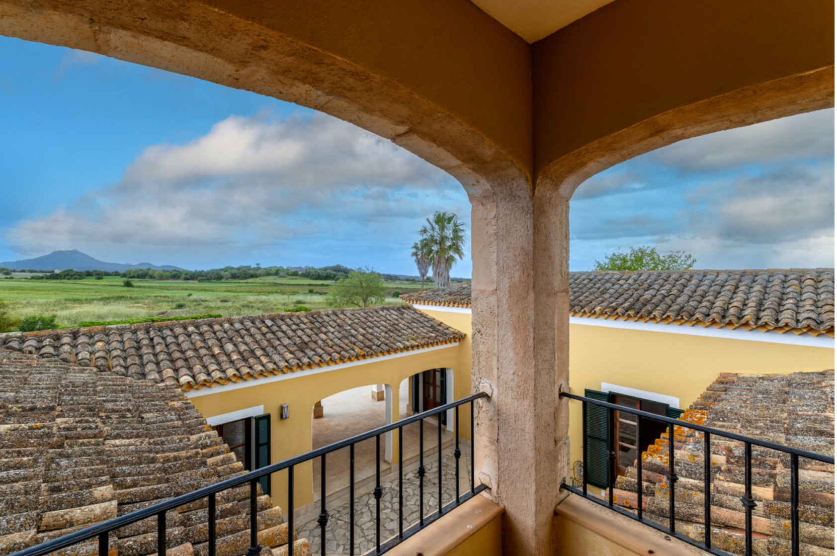 Scenic balcony view of Mediterranean countryside with tiled roofs and mountains in the distance under blue sky.