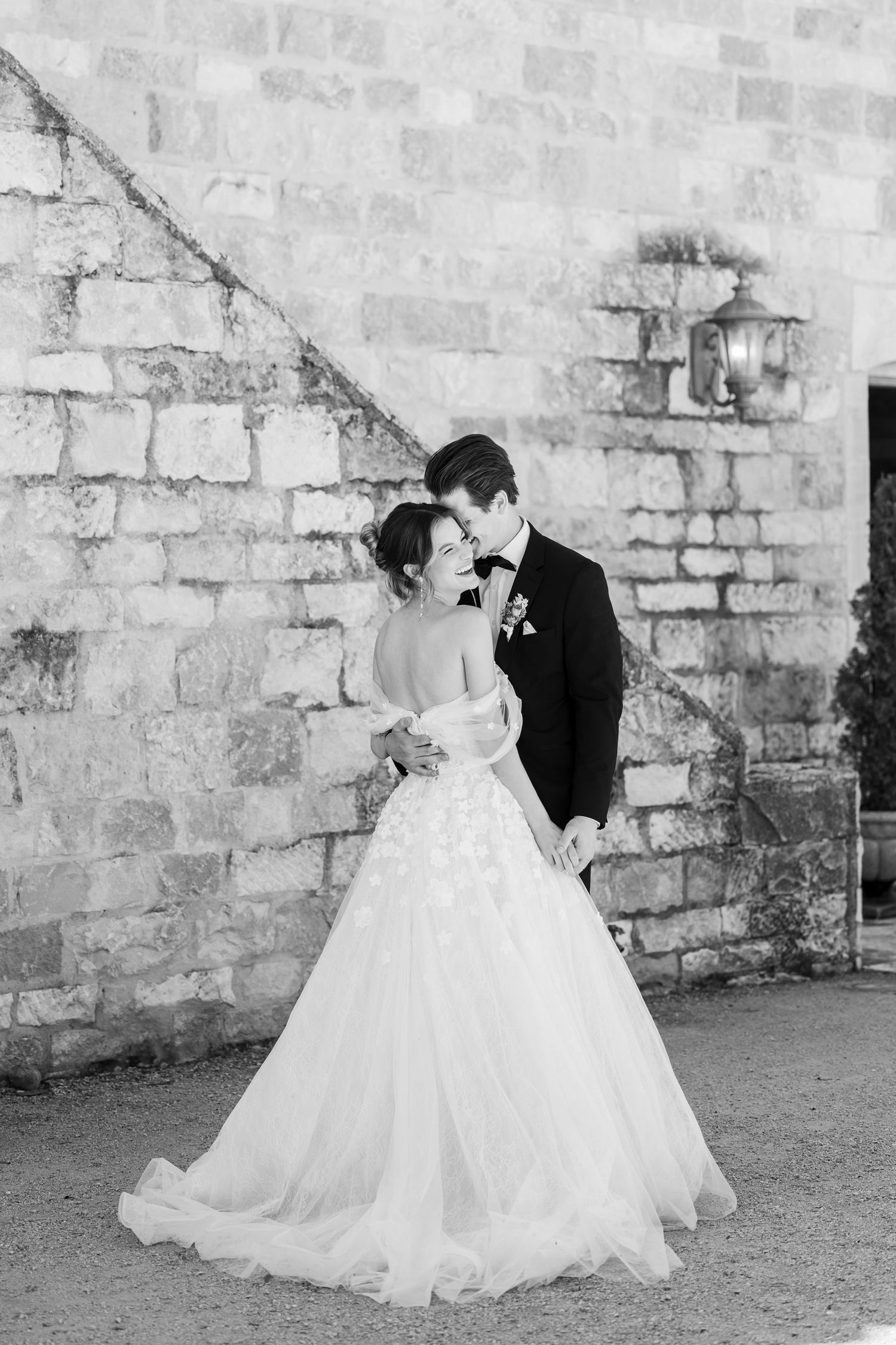 Wedding party in formal attire poses together outdoors with palm trees and brick building in background.