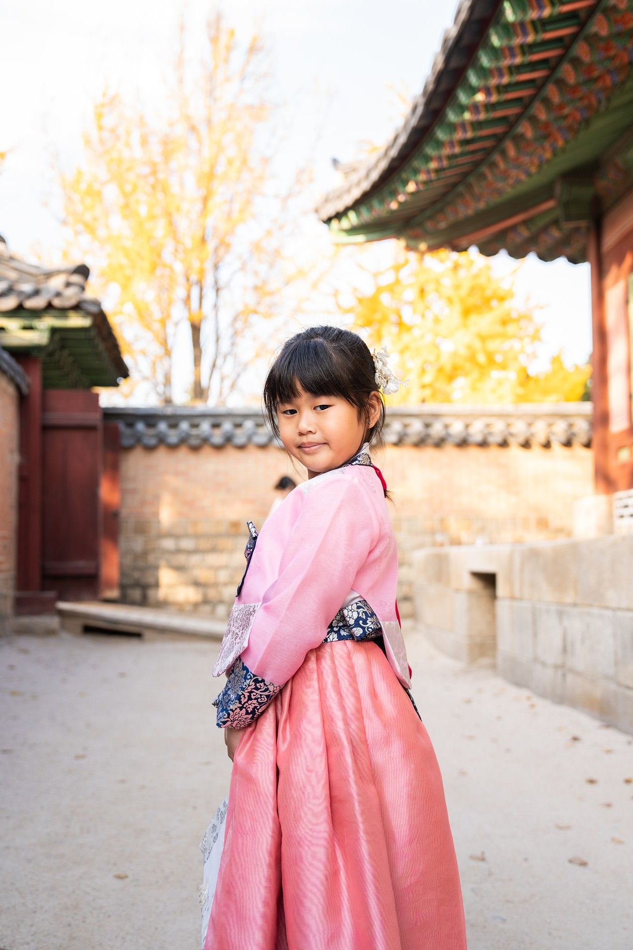 Traditional pink hanbok dress worn at historic Korean temple courtyard with autumn foliage and ornate architecture.