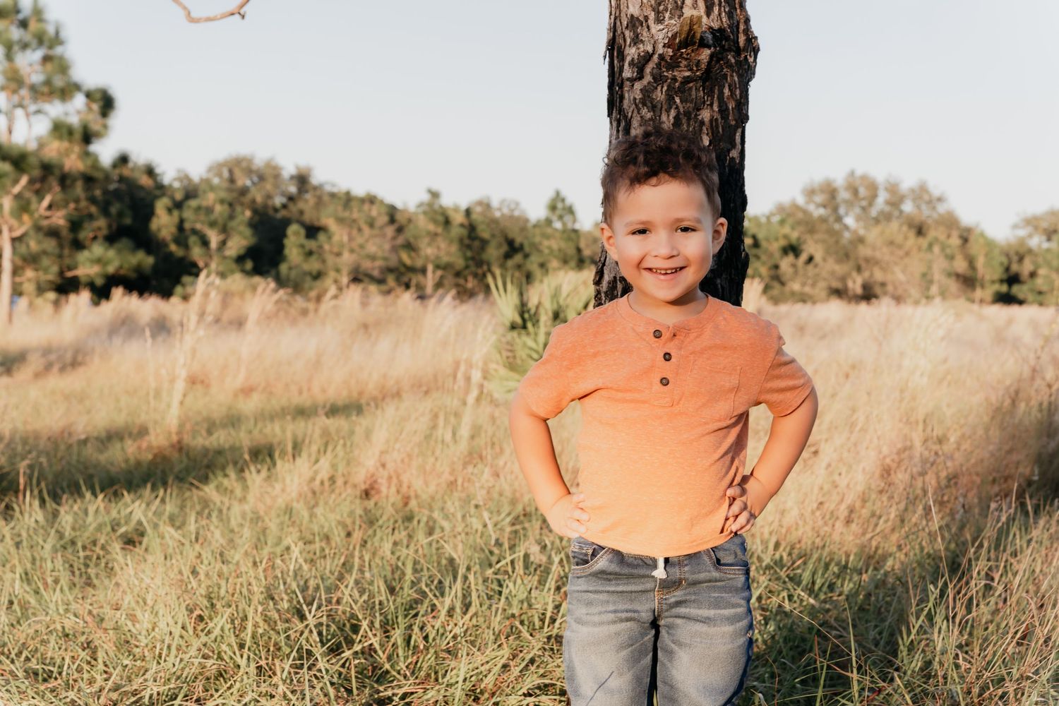 A child wearing an orange shirt and jeans stands in a golden field at sunset.