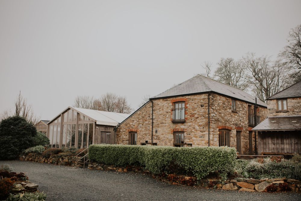 Charming stone farmhouse wedding venue with greenhouse and gravel driveway on an overcast day.