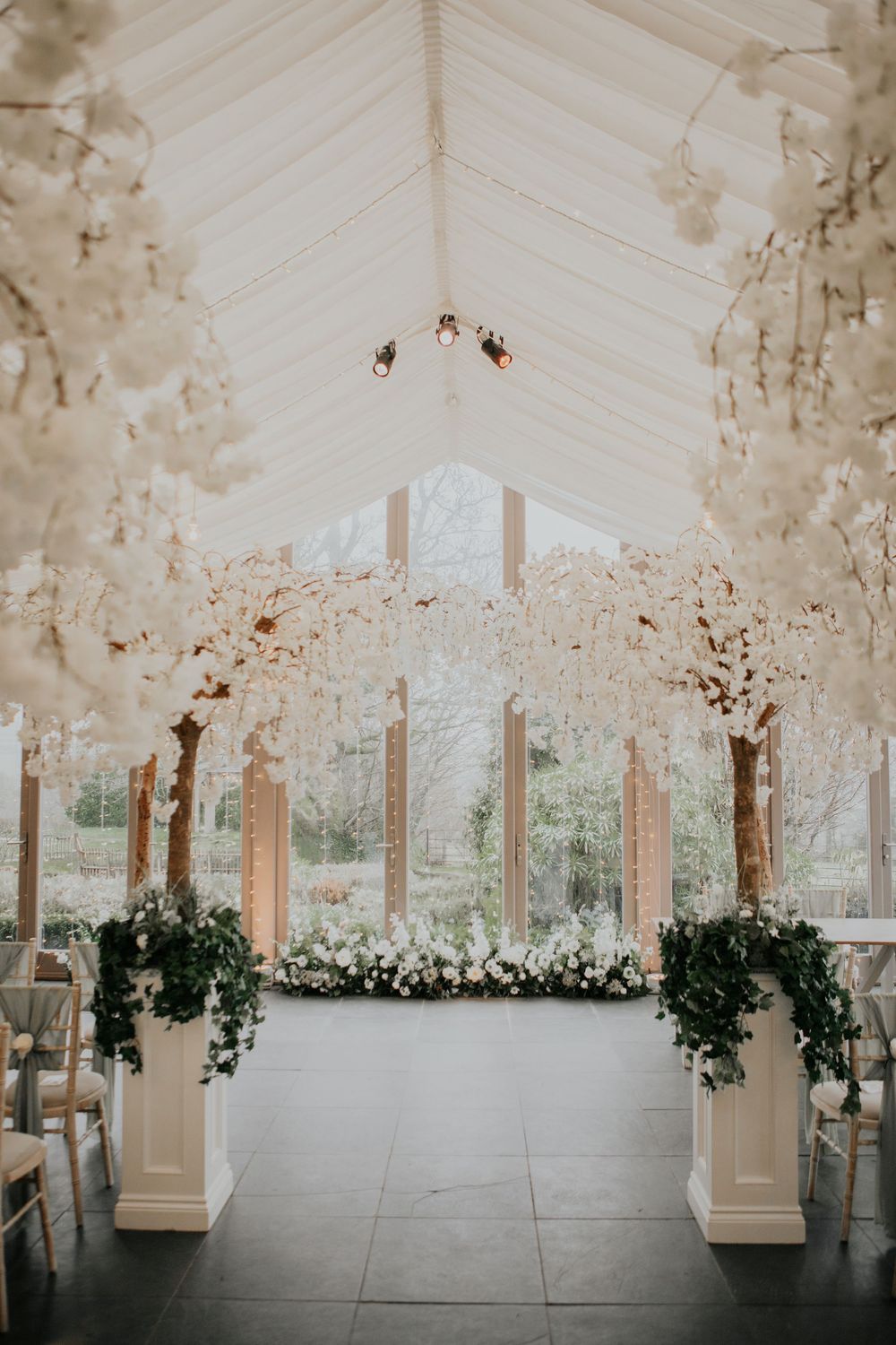 Elegant wedding ceremony space with white blossom trees, floral aisle, and glass ceiling venue.
