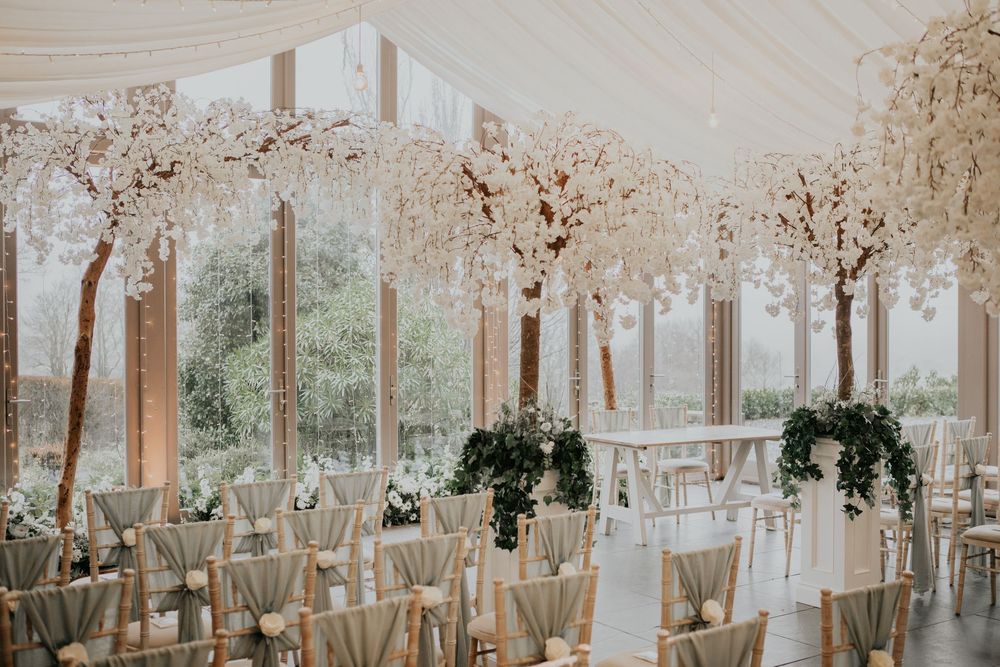Stunning wedding ceremony hall with tall white blossom trees and sage green draped chiavari chairs.
