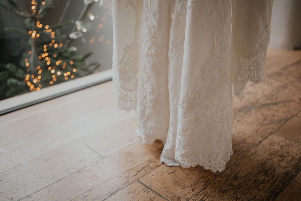 Close-up of intricate lace wedding dress hem on wooden floor with bokeh fairy lights.