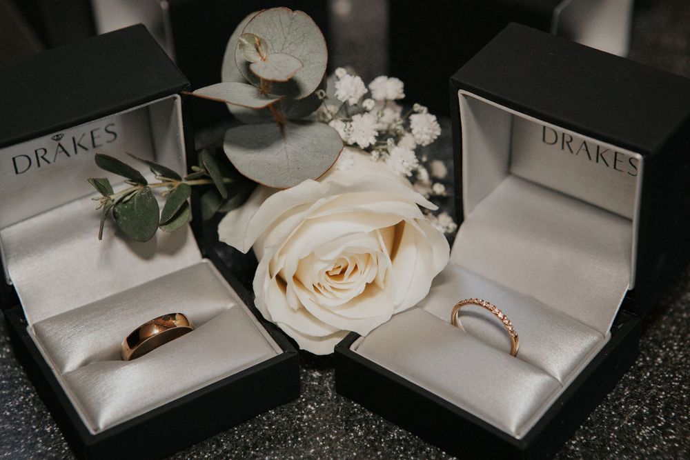 Two Drakes wedding rings in black boxes beside a white rose and eucalyptus.