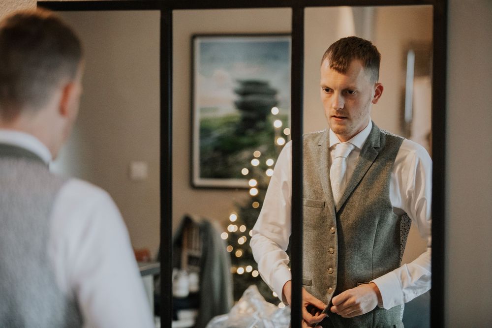 Groom in grey waistcoat adjusting cufflinks while looking in mirror.