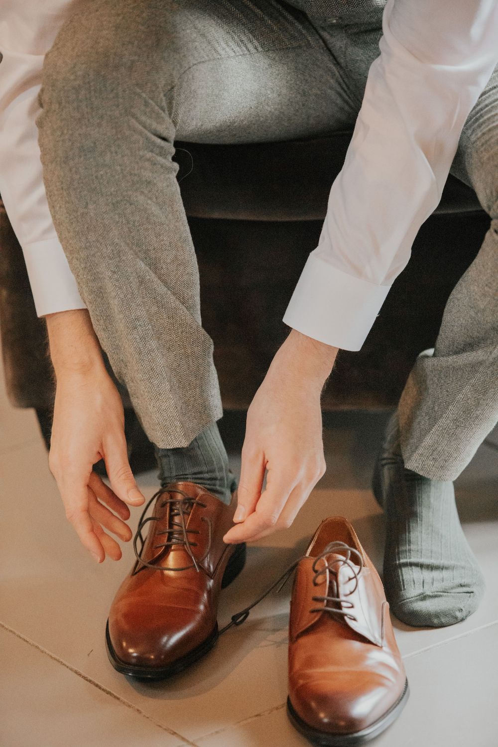 Groom tying laces on brown leather Oxford wedding shoes while seated.