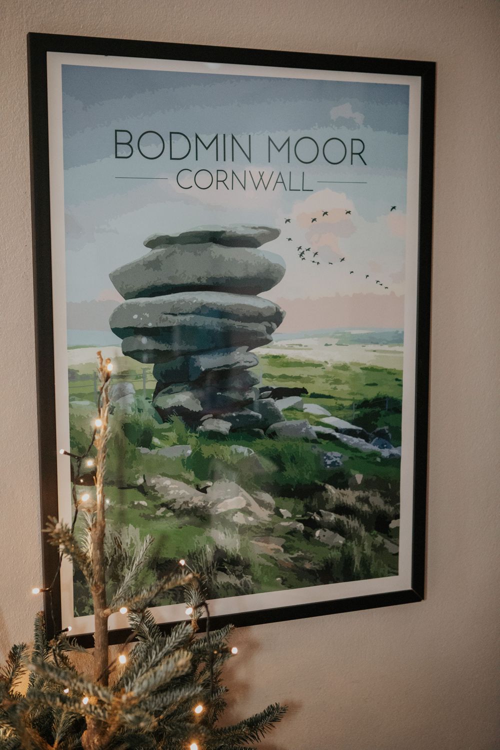Framed Bodmin Moor Cornwall travel poster on wall above fairy-lit tree.