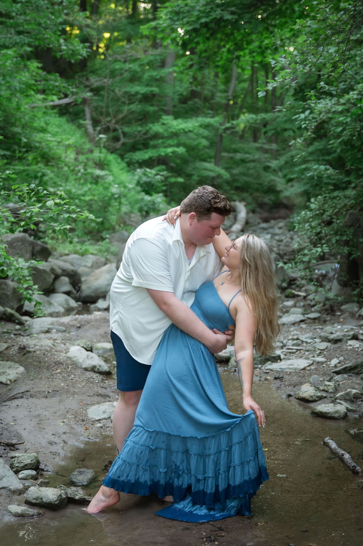 A romantic couple shares tender moments while standing in a shallow creek surrounded by lush green forest.