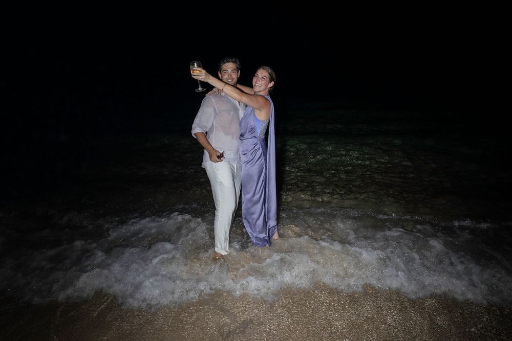 Two people in light colored clothing stand together in shallow ocean waves at night.