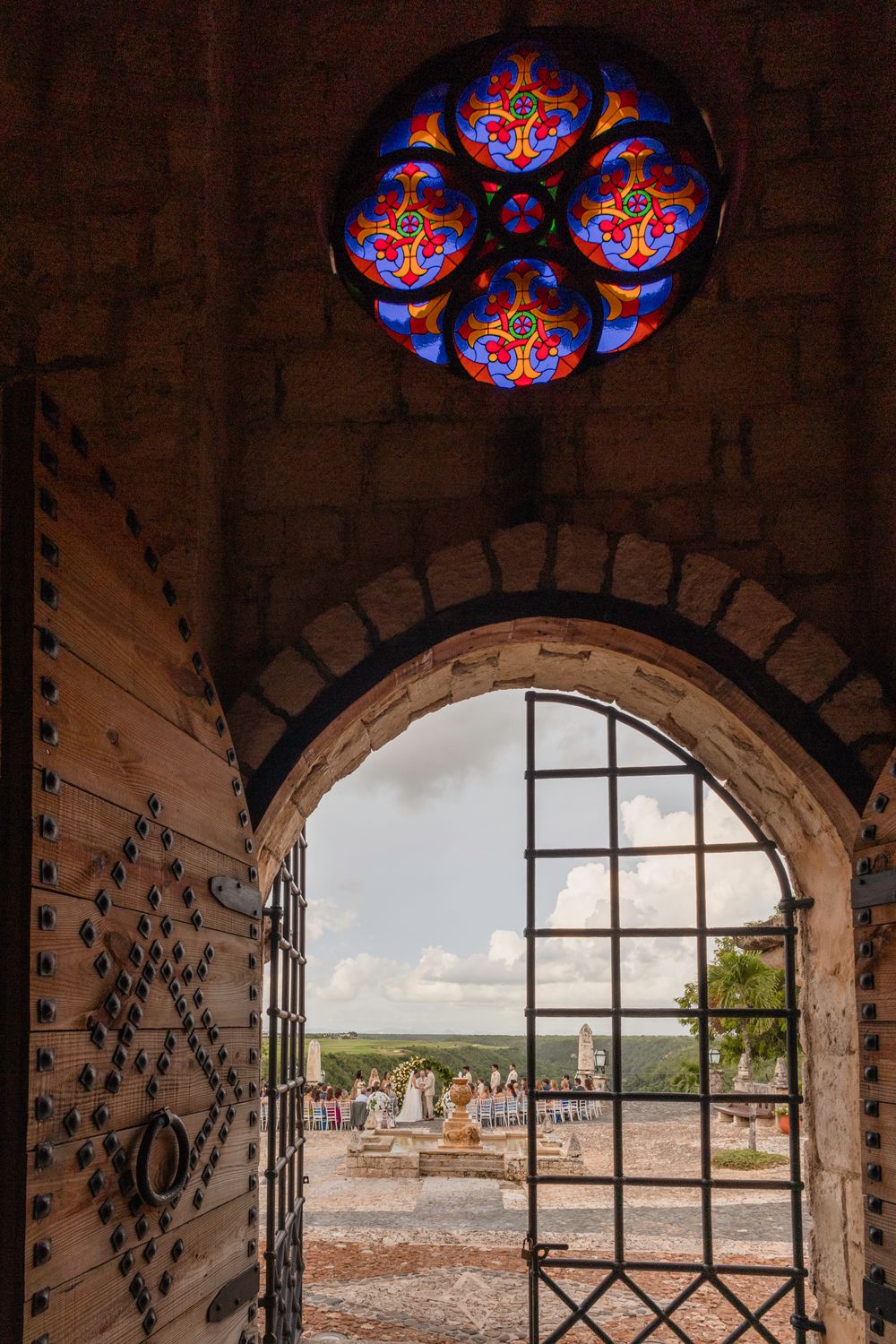 Ornate stained glass windows frame a view of a historic city through an arched stone window opening.