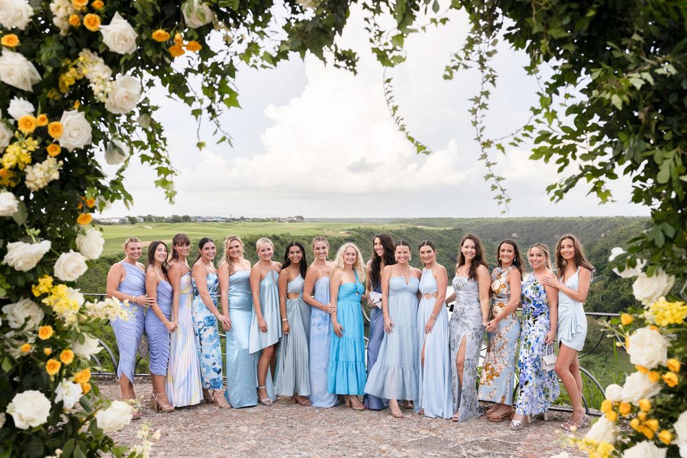 Wedding party in pale yellow and blue attire poses together under a floral arch with mountain views.