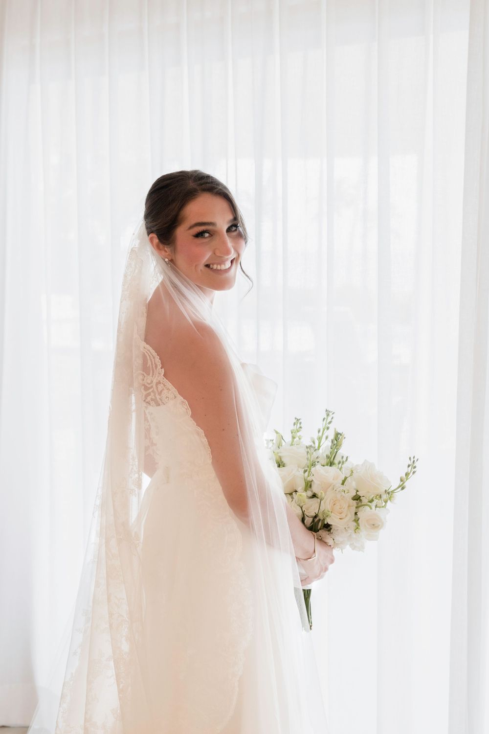 A bride in a flowing white gown and veil holds a white floral bouquet against a bright window backdrop.