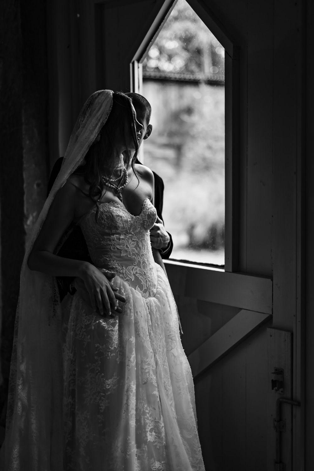 A bride in a lace wedding gown gazes thoughtfully through a window in dramatic black and white lighting.