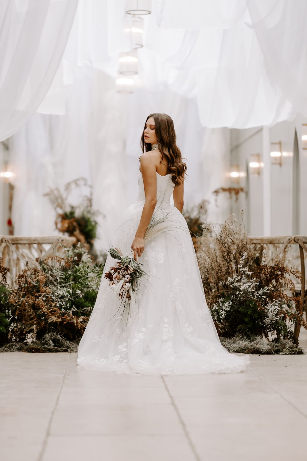 Bride in elegant white lace wedding gown poses in garden setting with flowing veil and dried floral bouquet.