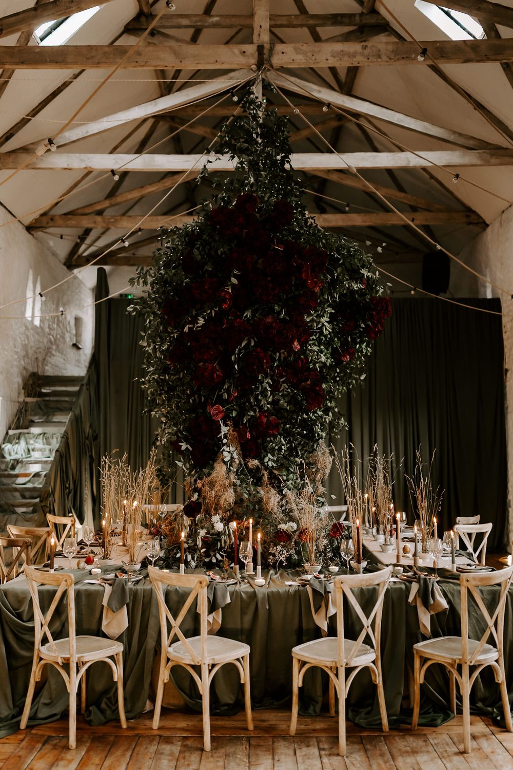Rustic barn wedding reception setup with dark green drapes, wooden cross-back chairs, and hanging floral arrangements.