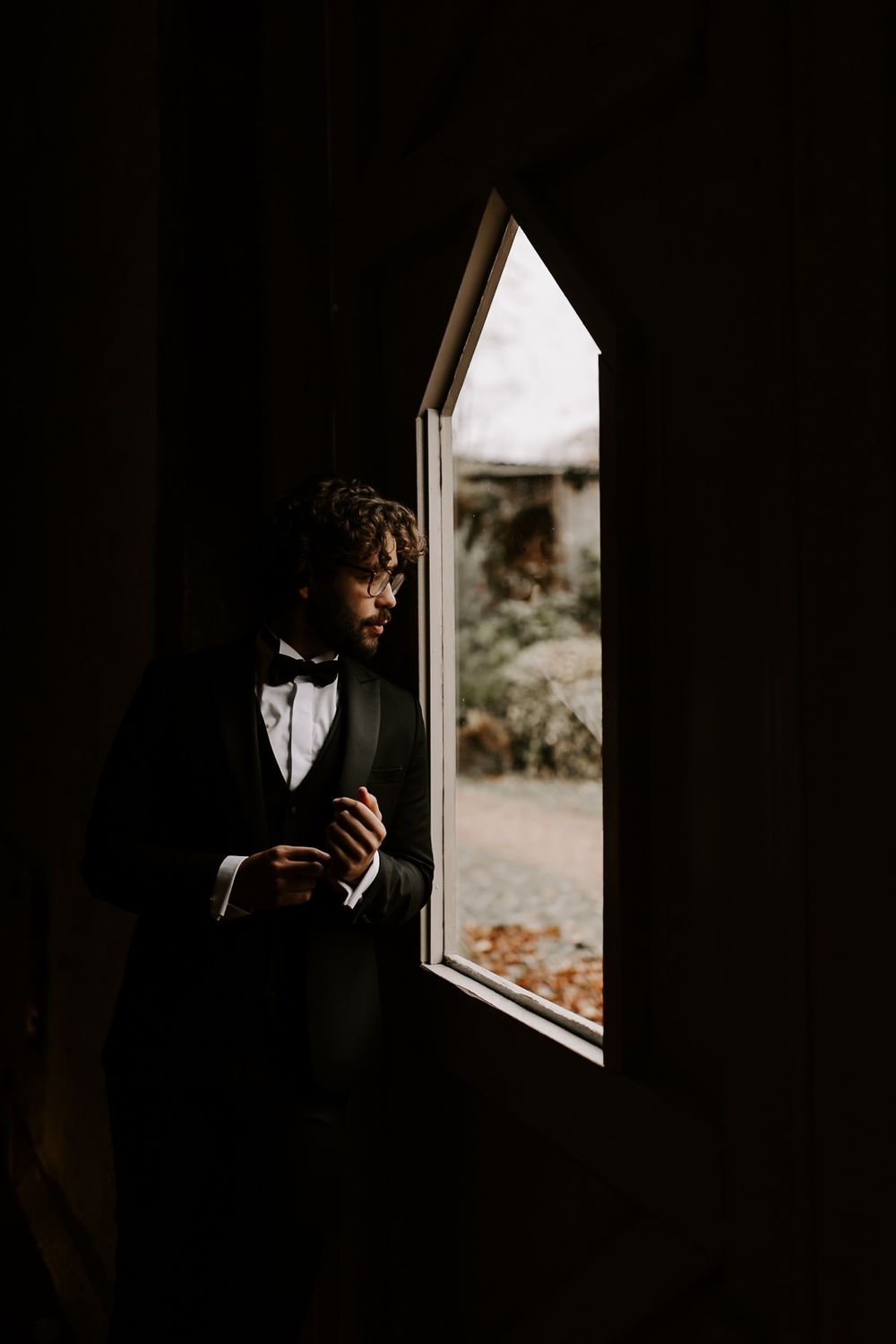 A dark silhouette stands by a window in a dimly lit room creating a moody atmospheric portrait series.