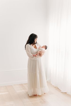 Séance fine art toute en lumière d’une nouvelle maman tenant son nouveau-né contre elle dans une robe longue et vaporeuse