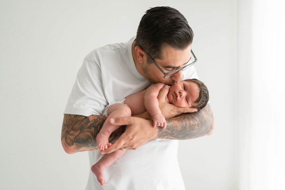 Papa tenant son nouveau-né dans ses bras et déposant un baiser sur sa joue lors d'un photoshoot nouveau-né en studio de Saint-Augustin