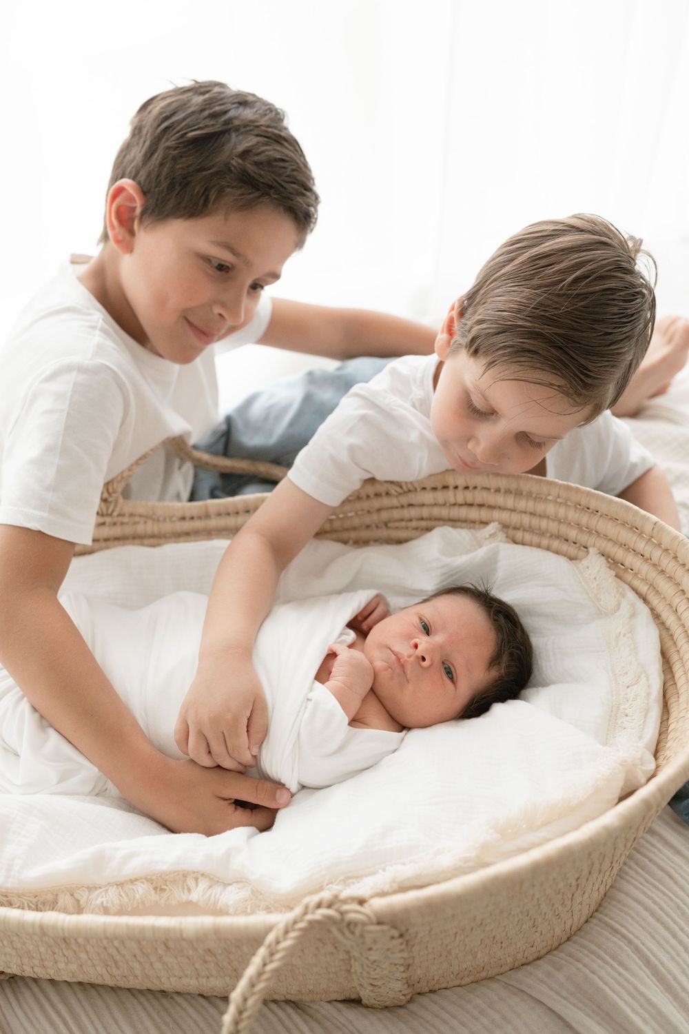 Grands frères penchés sur le panier en osier dans lequel leur nouveau petit frère se repose lors de leur séance photo nouveau-né près Québec