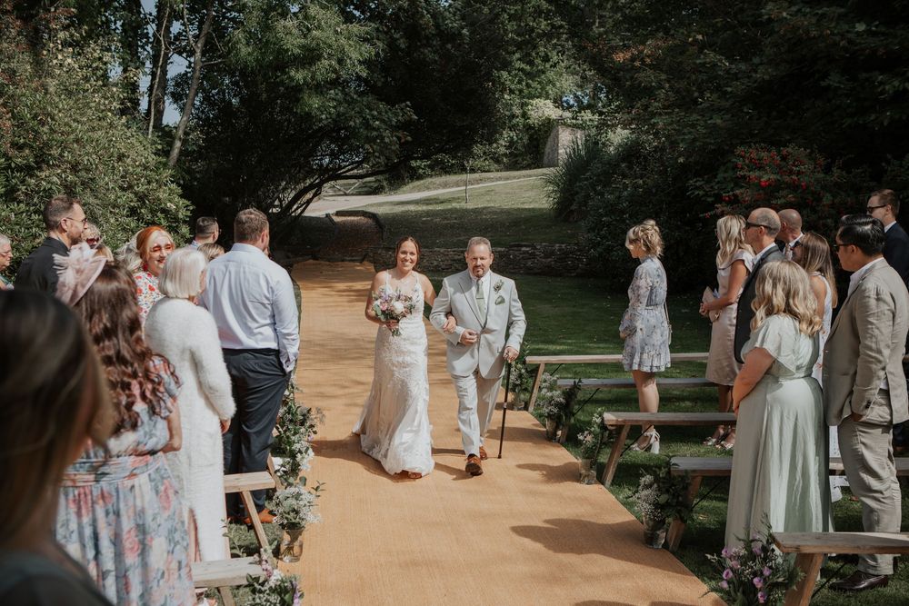 Bride walking down the outdoor aisle with her father past seated wedding guests in a garden ceremony.