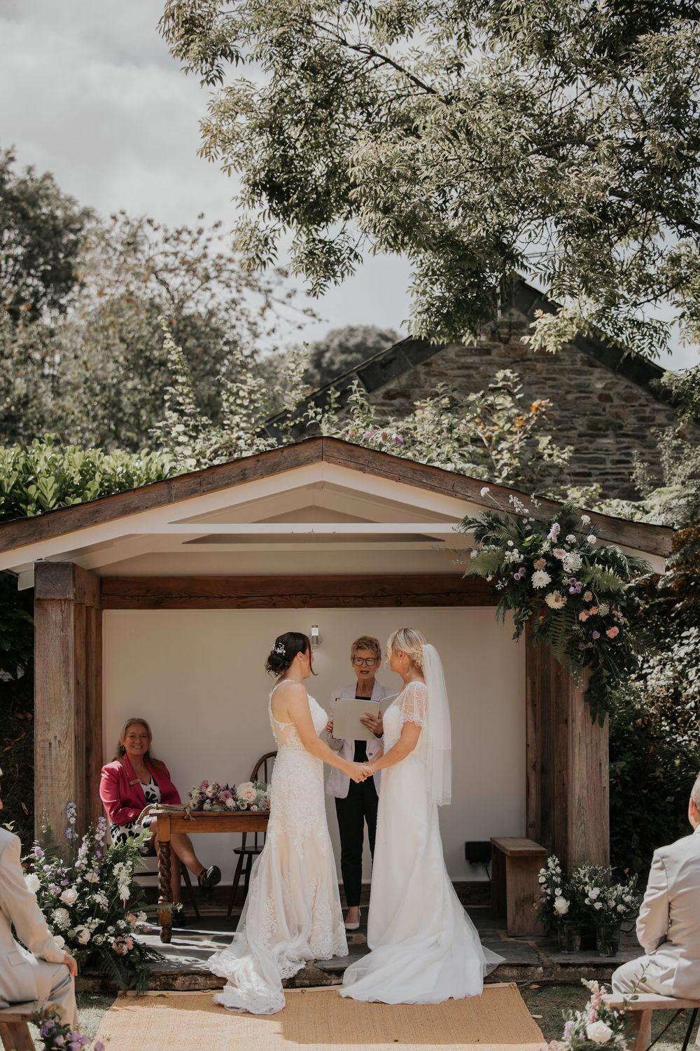 Two brides exchanging vows at an outdoor wedding ceremony beneath a floral-adorned wooden altar.