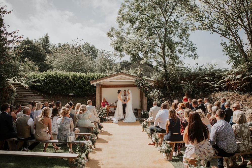 Two brides at the altar during an outdoor garden wedding ceremony with guests seated on wooden benches.