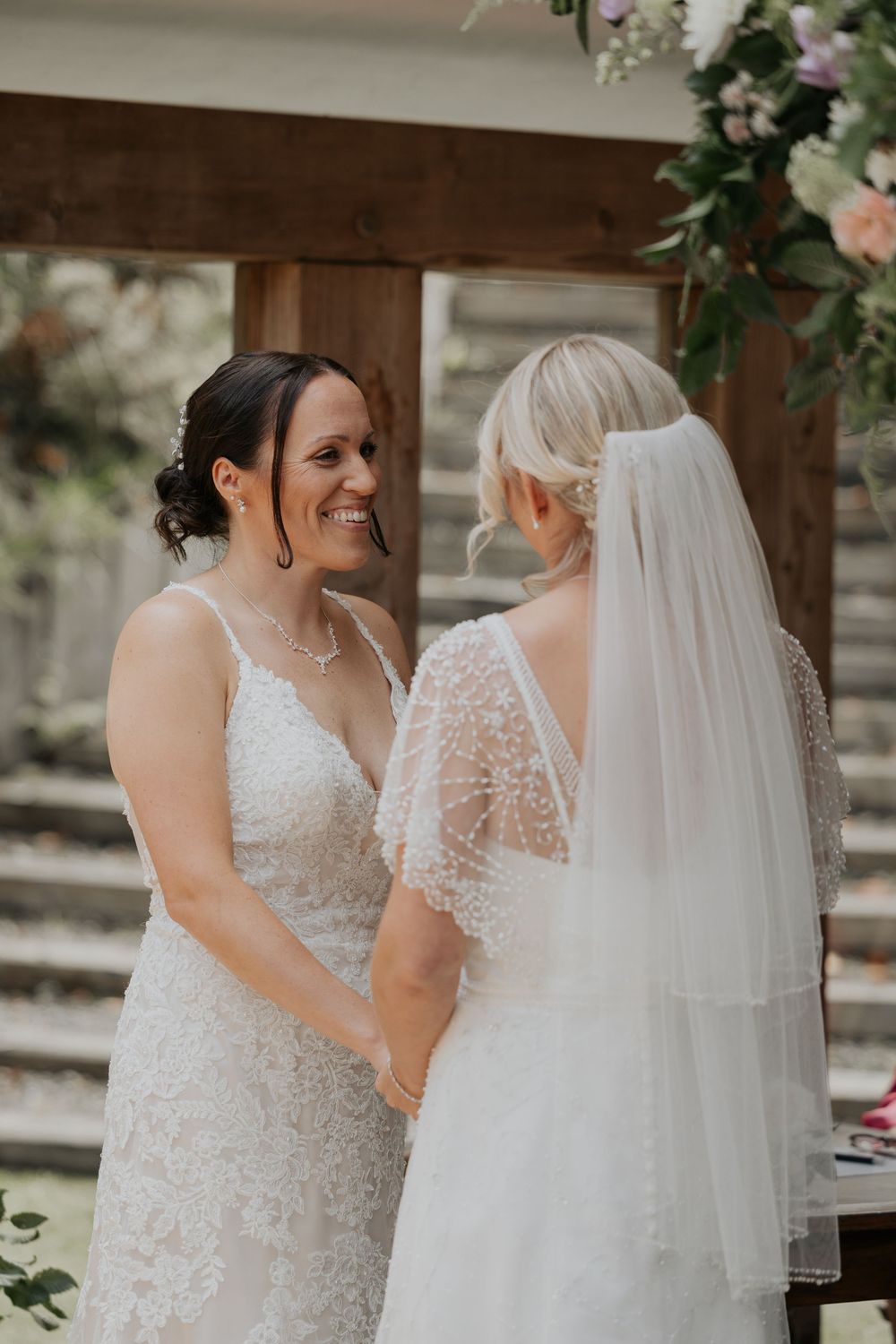 Two brides holding hands and smiling at each other during their outdoor wedding ceremony.