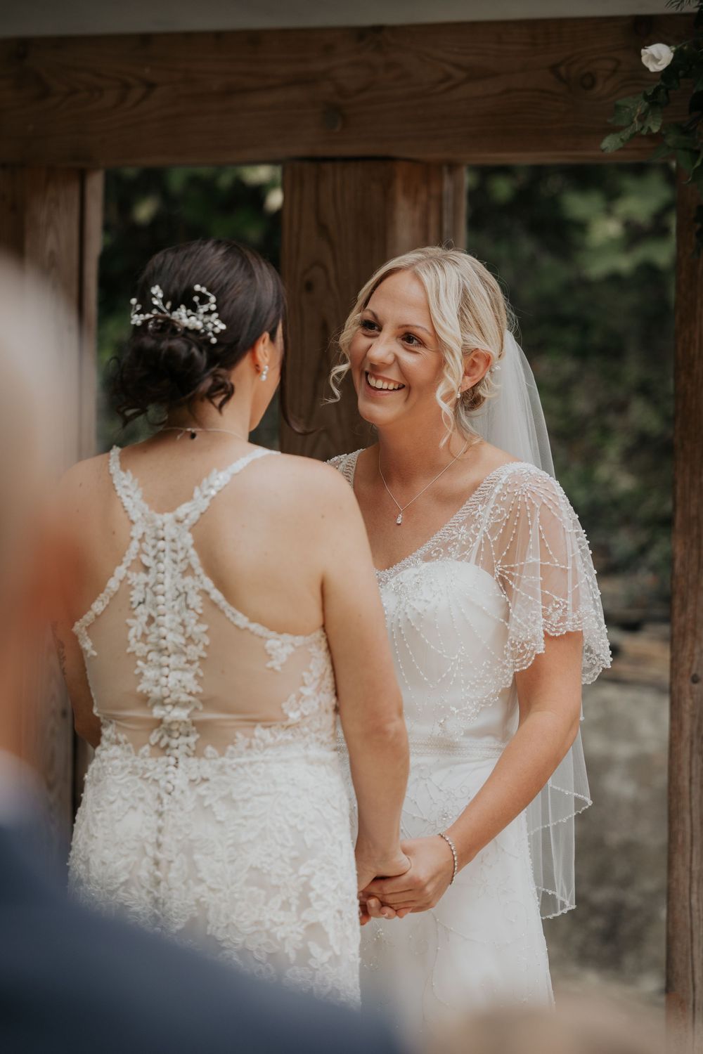 Two brides in white lace wedding dresses hold hands and smile at each other during their wedding ceremony.