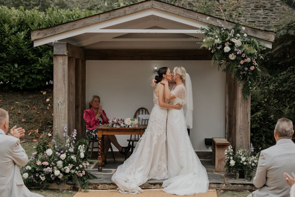 Two brides share a first kiss under a floral-decorated wooden arbor during their outdoor wedding ceremony.