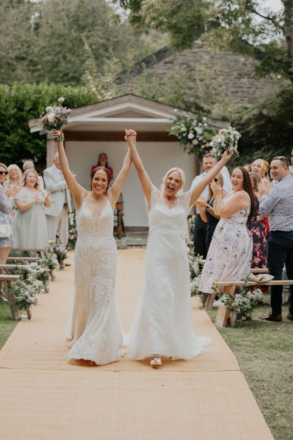 Two brides celebrate joyfully raising bouquets while walking down the aisle after their wedding ceremony.