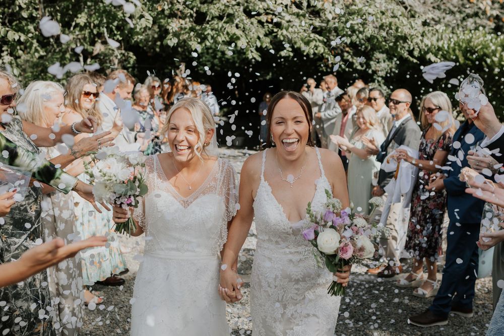 Two brides laugh as guests shower them with white confetti during their outdoor wedding celebration.