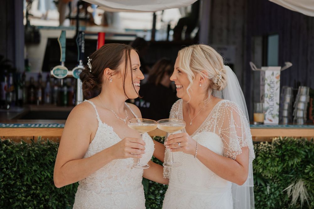Two brides in white lace gowns smile and clink cocktail glasses together at their wedding reception bar.