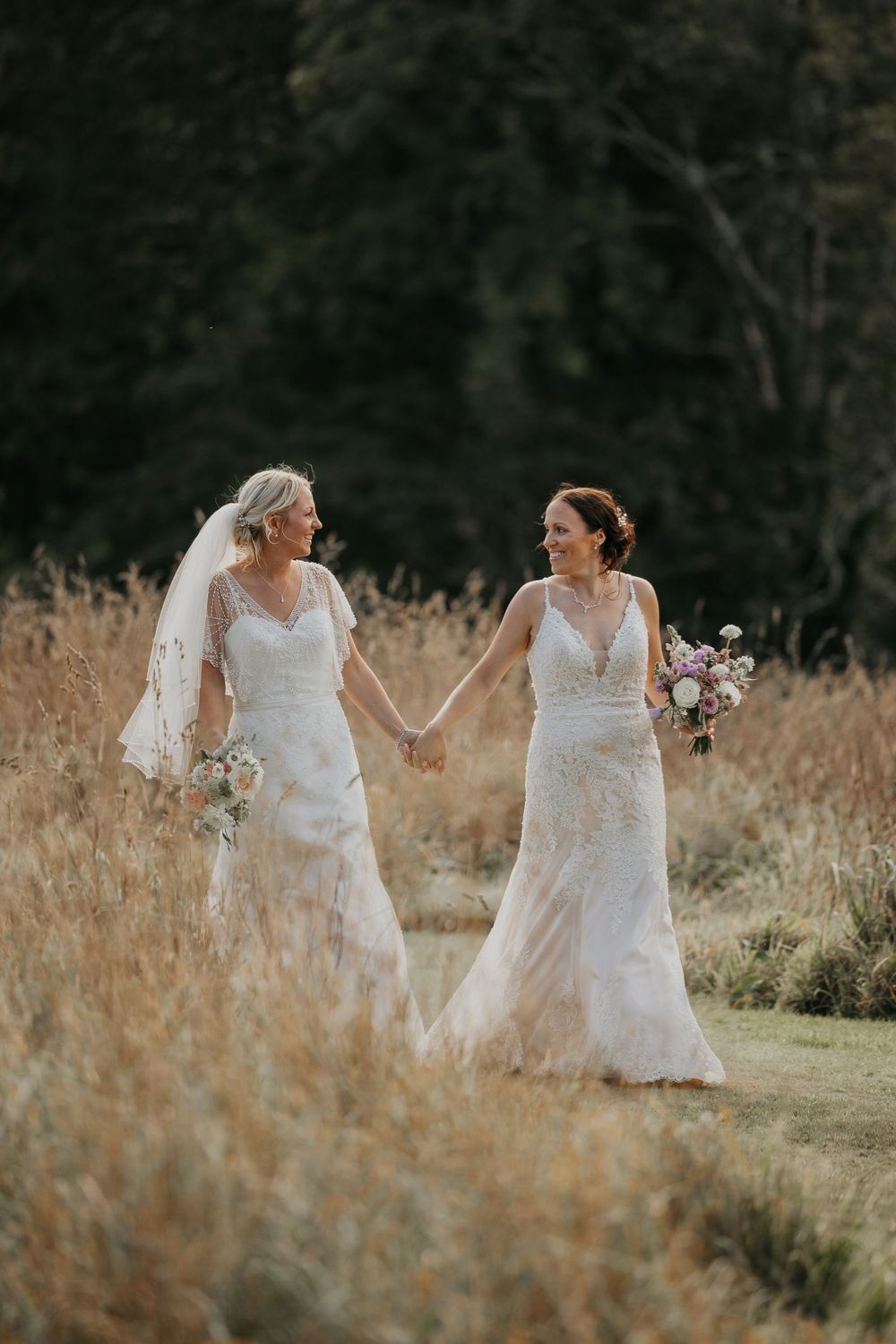 Two brides in white lace wedding gowns hold hands while walking through a golden meadow on their wedding day.