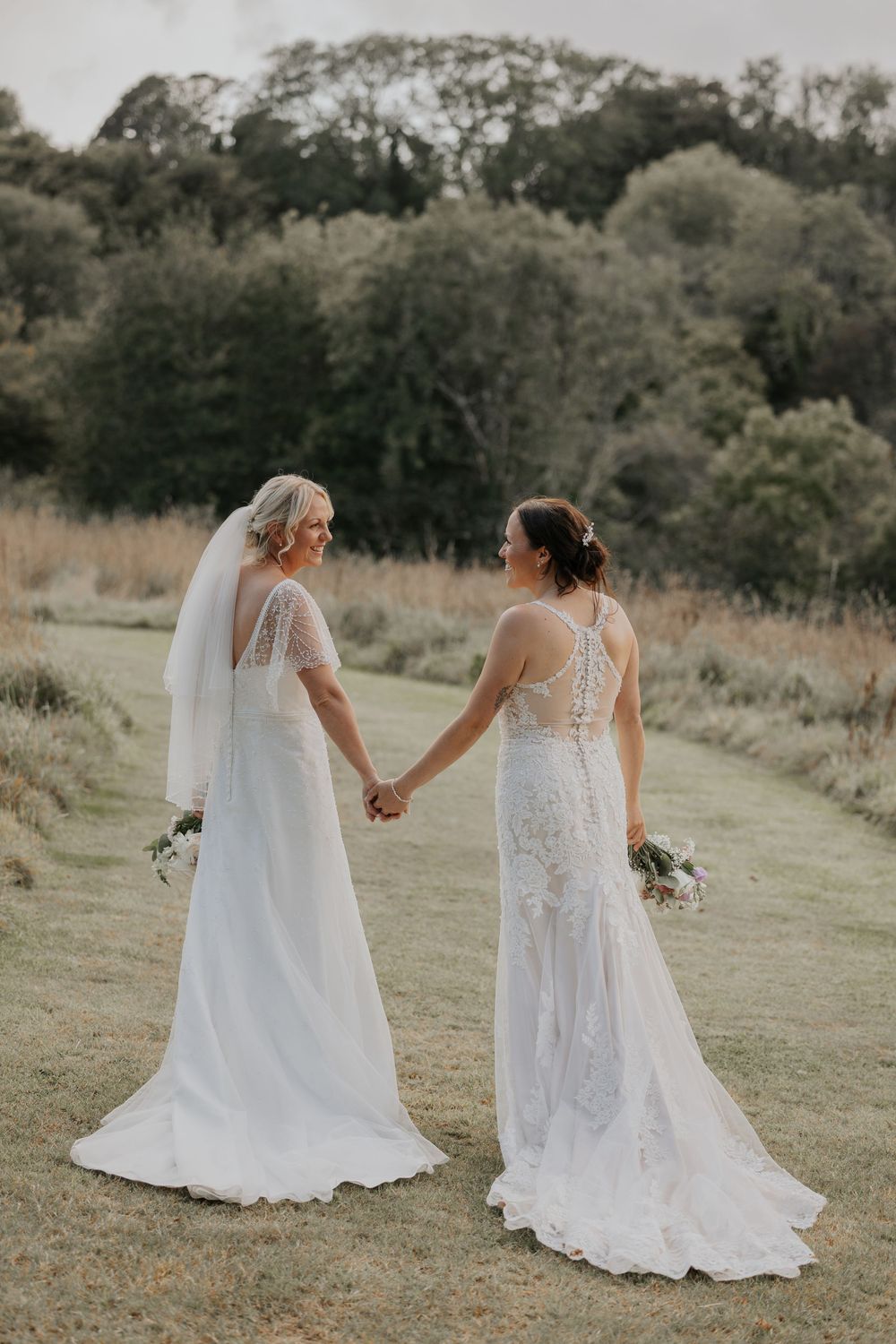 Two brides in white wedding dresses hold hands while walking through a lush green countryside landscape.