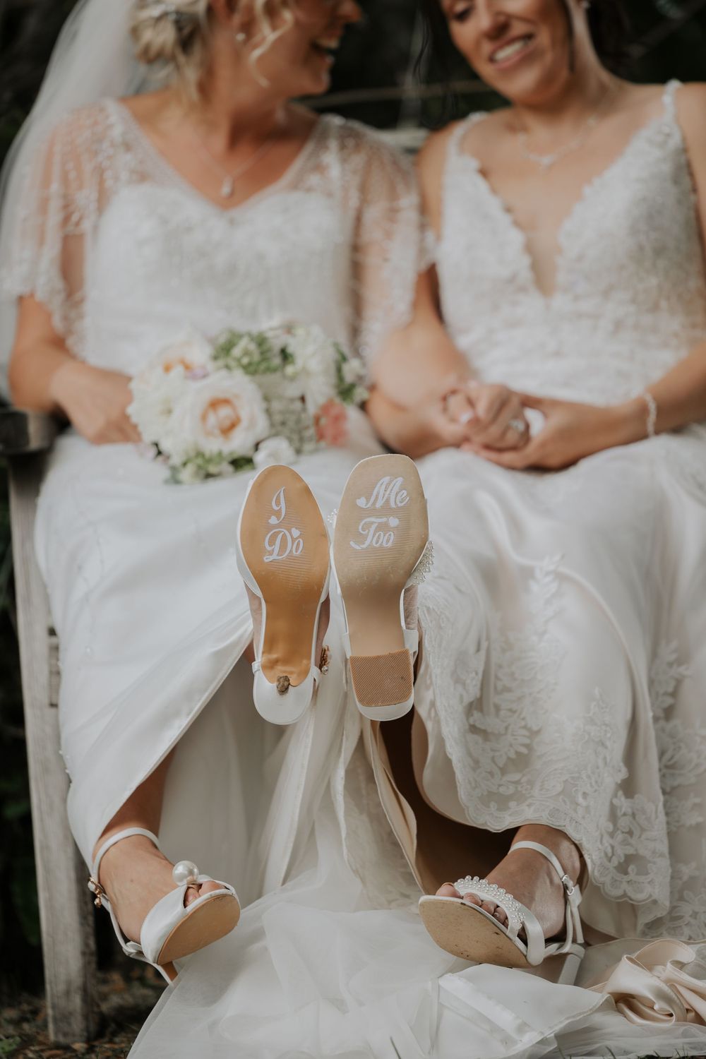 Wedding shoes with I Do and Me Too written on soles as two brides sit together holding bouquets.