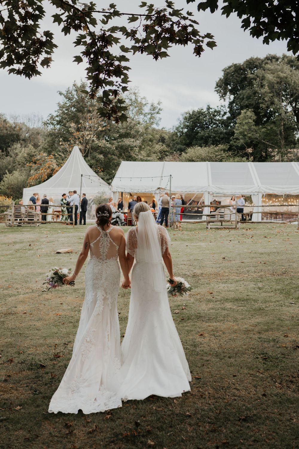 Two brides in white gowns walk hand in hand toward a marquee tent at their outdoor garden wedding.