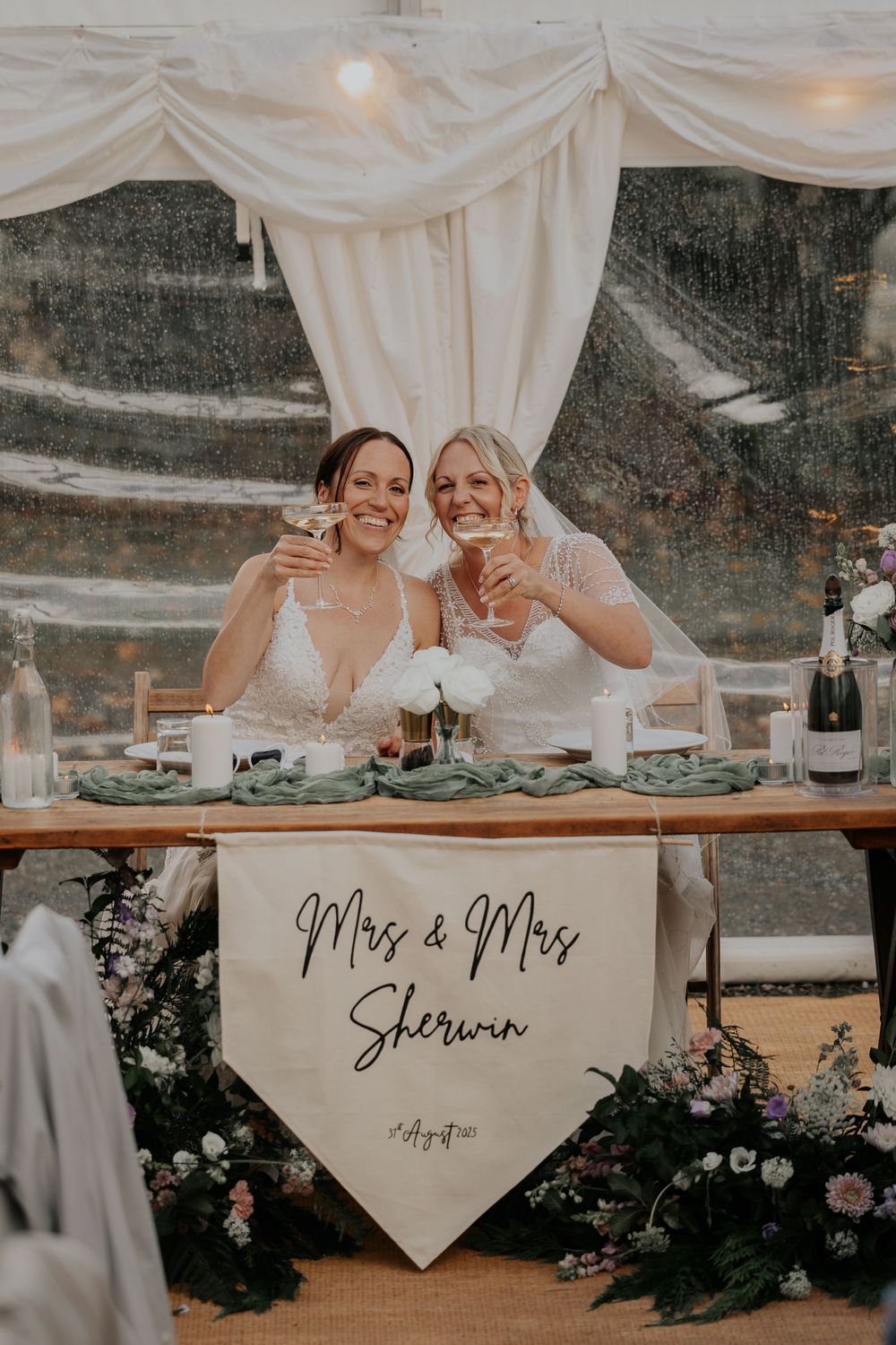 Two brides toast champagne at their reception sweetheart table with Mrs & Mrs Sherwin banner displayed.
