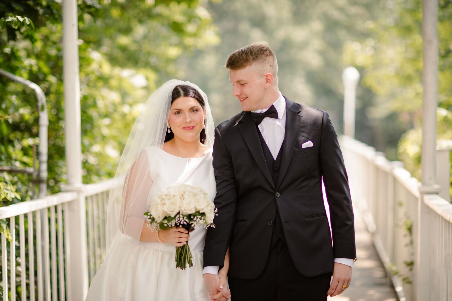 A couple walks hand in hand across a rustic wooden bridge surrounded by lush green forest scenery.